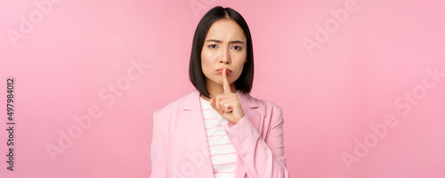 Hush, taboo concept. Portrait of asian businesswoman showing shush gesture, shhh sign, press finger to lips, standing over pink background in suit photo