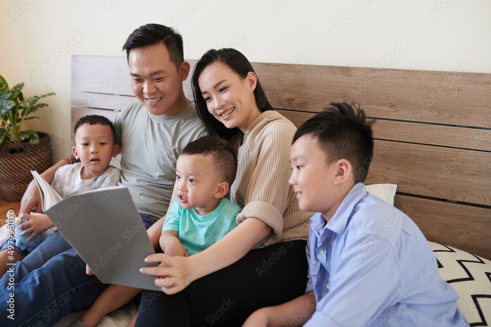 Happy parents and three children sitting on bed and reading new book together Happy parents and three children sitting on bed and reading new book together