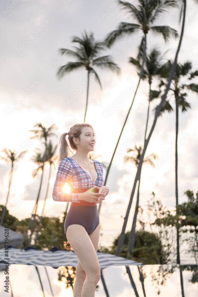 Woman in one piece swimsuit standing near swimming pool, sunset moment.