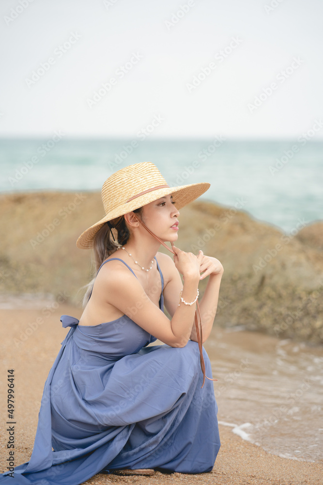 Woman wearing blue dress and straw hat sitting on the beach.