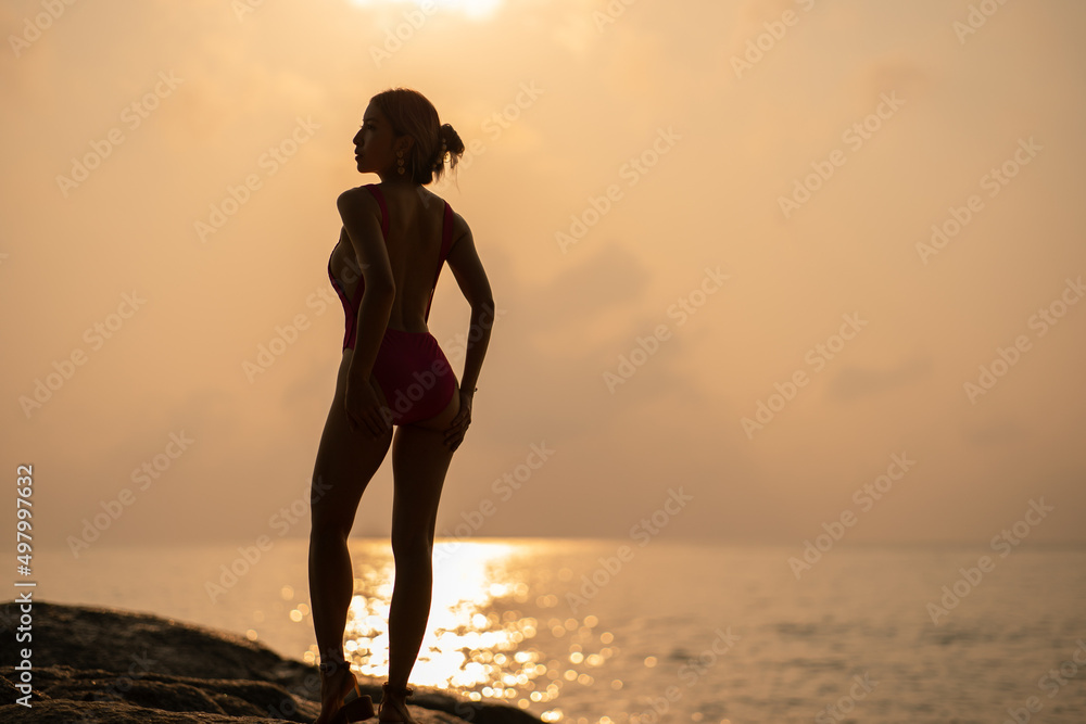 silhouette : Woman wearing pink one piece swimsuit enjoy romantic sunset moment on the stone, tropical beach.