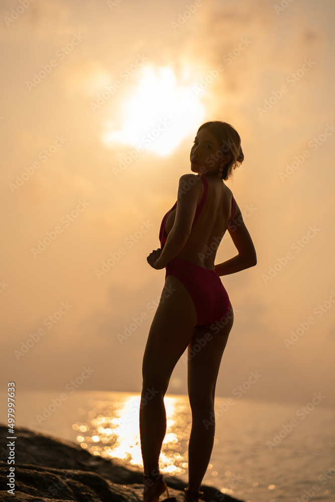 silhouette : Woman wearing pink one piece swimsuit enjoy romantic sunset moment on the stone, tropical beach.