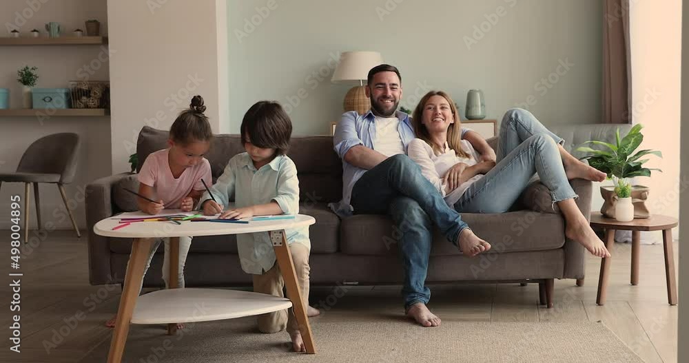 Young beautiful couple relaxing sit on couch, their preschoolers kids drawing pictures in paper sketchbook with colored pencils, spend leisure at modern cozy living room. Family weekend, hobby concept Young beautiful couple relaxing sit on couch, their preschoolers kids drawing pictures in paper sketchbook with colored pencils, spend leisure at modern cozy living room. Family weekend, hobby concept
