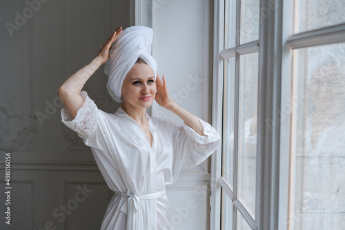 Young woman wrapped in towel and white robe standing next to bedroom window drying her hair after a morning shower. Smiling lady with towel on her head looking at empty space, home interior, panorama