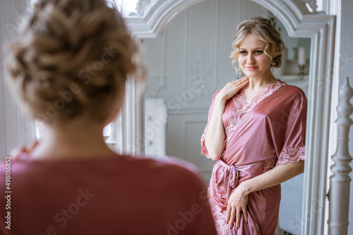 Happy woman looks at her reflection in mirror in pink silk robe near window in the morning. Beautiful makeup and hair. Skin and body care at home. Women's beauty and health