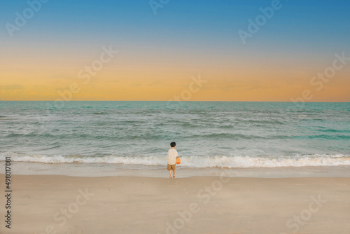 Back view of little boy standing on the beach looking at the sea, beautiful sky in evening.