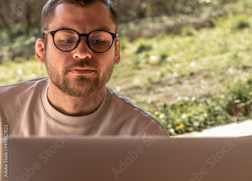 Handsome confident man with stubble on his face wearing glasses looking at laptop screen.