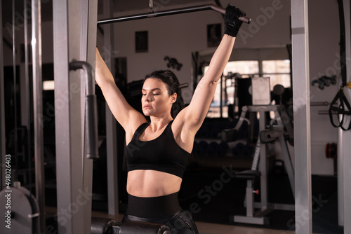 side view of a young beautiful fitness lady in black sportswear sits on a simulator and performs exercises for the arms and back in gym. concept of self-discipline and motivation.