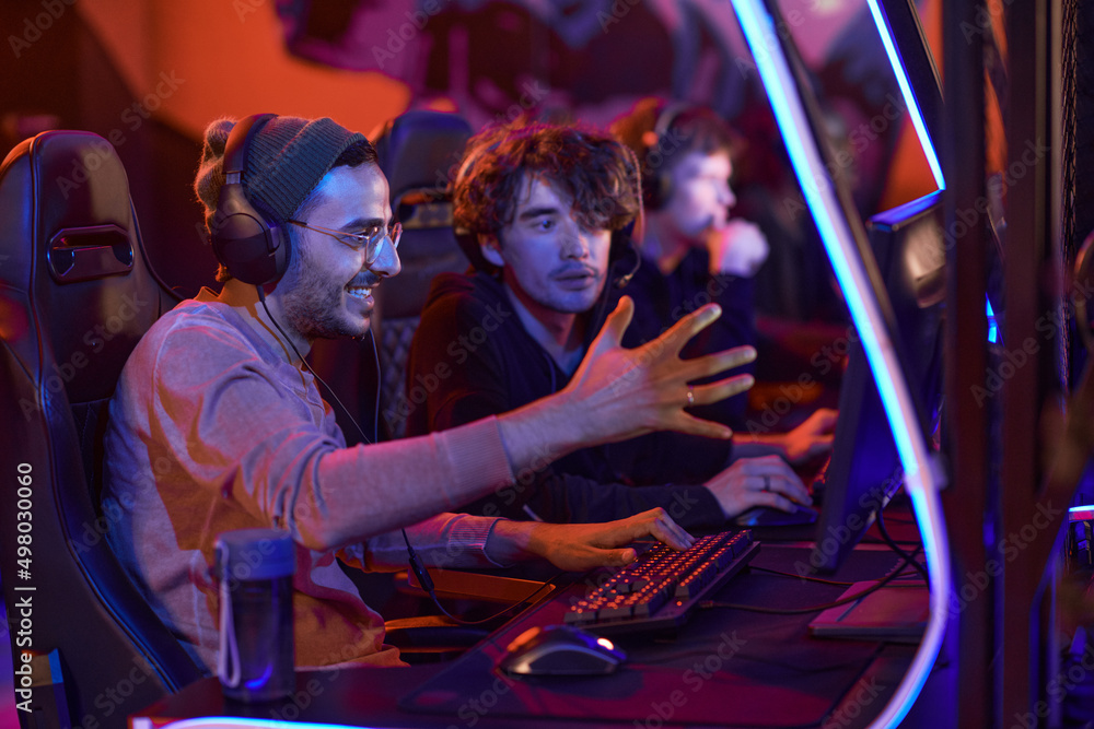 Positive young multi-ethnic guys in headsets sitting in front of computer and playing game together in esports bar