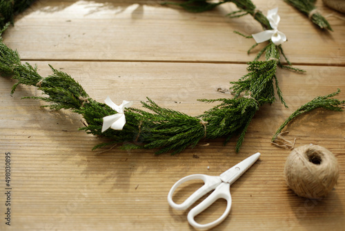 scissors, sisal twine and cedar leaves wreath tied up with white ribbon bows on wooden craft table. crafting outdoor. space for copy.