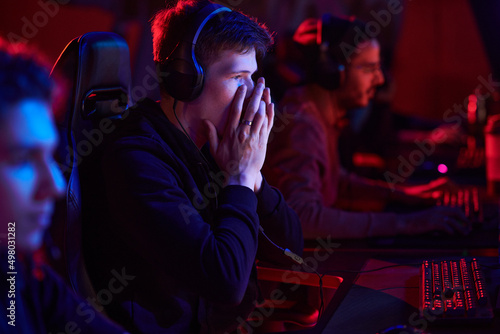 Shocked teenage gamer in headphones covering mouth and looking at monitor while losing game at esports tournament