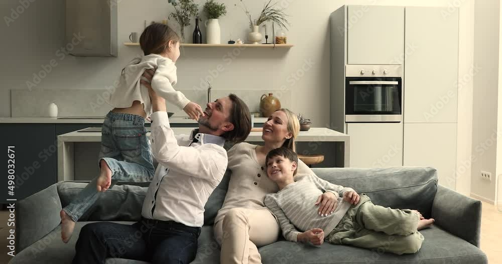 Happy family homeowners relax on sofa at cozy studio apartment play spend free time together. Smiling mother cuddle tween age son watching father tossing in air excited little preschool age daughter Happy family homeowners relax on sofa at cozy studio apartment play spend free time together. Smiling mother cuddle tween age son watching father tossing in air excited little preschool age daughter