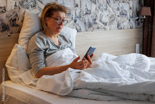 An elderly woman uses a mobile phone while lying in bed at home. A sick elderly woman uses a smartphone while lying in bed.