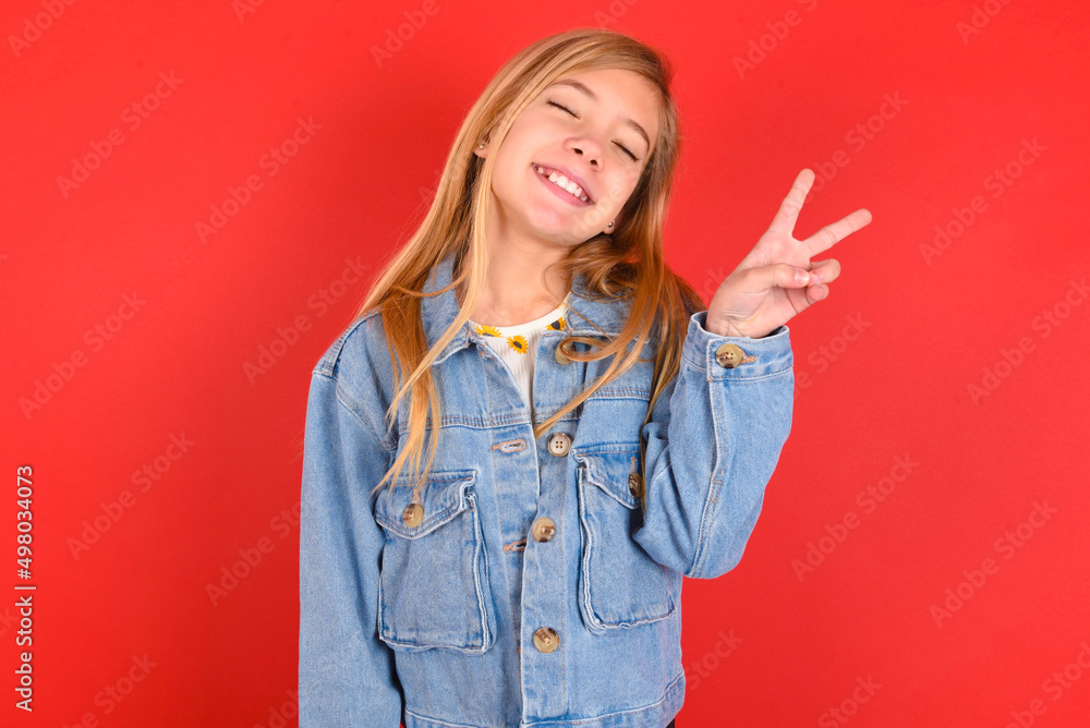 blonde little kid girl wearing denim jacket over red background smiling with happy face winking at the camera doing victory sign. Number two. blonde little kid girl wearing denim jacket over red background smiling with happy face winking at the camera doing victory sign. Number two.
