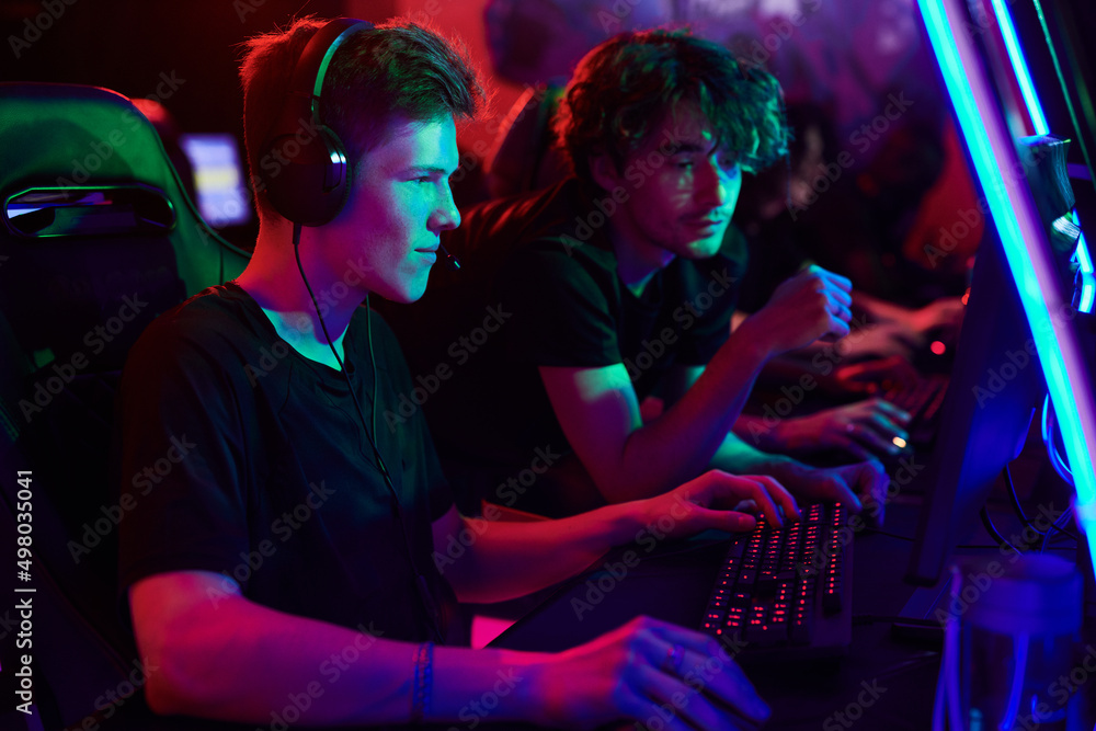 Curious young man in headphones playing online computer game under control of cybersport coach in gaming club Curious young man in headphones playing online computer game under control of cybersport coach in gaming club