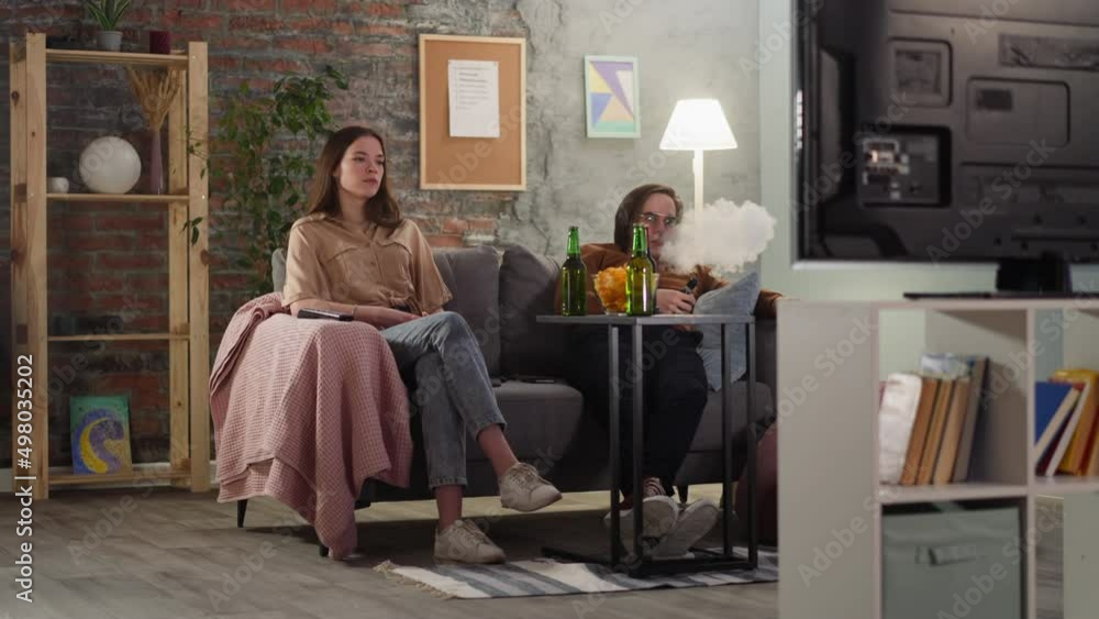 Man with glasses smokes vape sitting with tranquil young woman at football game broadcasting in apartment. Sports fans at awkward dating at home Man with glasses smokes vape sitting with tranquil young woman at football game broadcasting in apartment. Sports fans at awkward dating at home