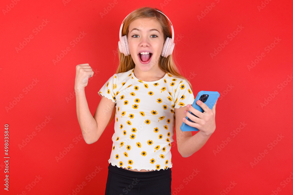 Positive blonde little kid girl wearing daisy t-shirt red background holds modern cell phone connected to headphones, clenches fist from good emotions, exclaims with joy, Positive blonde little kid girl wearing daisy t-shirt red background holds modern cell phone connected to headphones, clenches fist from good emotions, exclaims with joy,
