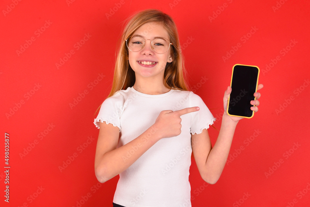Smiling blonde little kid girl wearing denim jacket over red background Mock up copy space. Pointing index finger on mobile phone with blank empty screen Smiling blonde little kid girl wearing denim jacket over red background Mock up copy space. Pointing index finger on mobile phone with blank empty screen