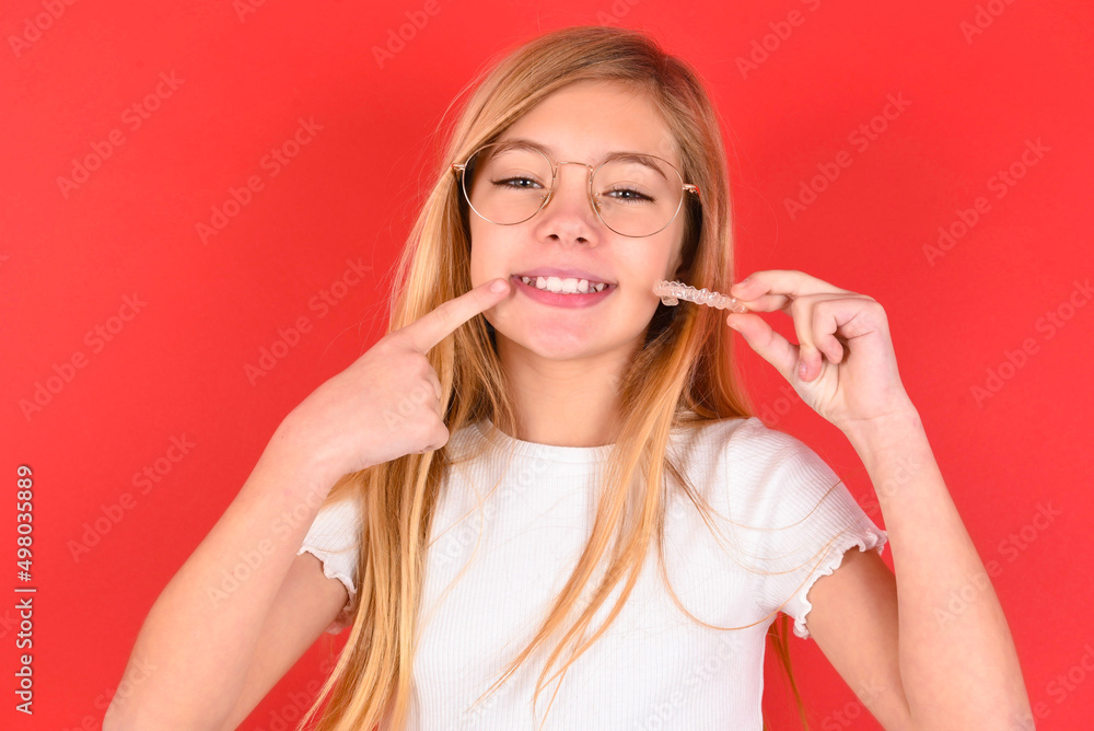 blonde little kid girl wearing denim jacket over red background holding an invisible aligner and pointing to her perfect straight teeth. Dental healthcare and confidence concept. blonde little kid girl wearing denim jacket over red background holding an invisible aligner and pointing to her perfect straight teeth. Dental healthcare and confidence concept.