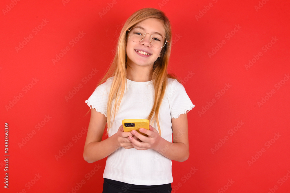 blonde little kid girl wearing white t-shirt over red background Mock up copy space. Using mobile phone, typing sms message blonde little kid girl wearing white t-shirt over red background Mock up copy space. Using mobile phone, typing sms message
