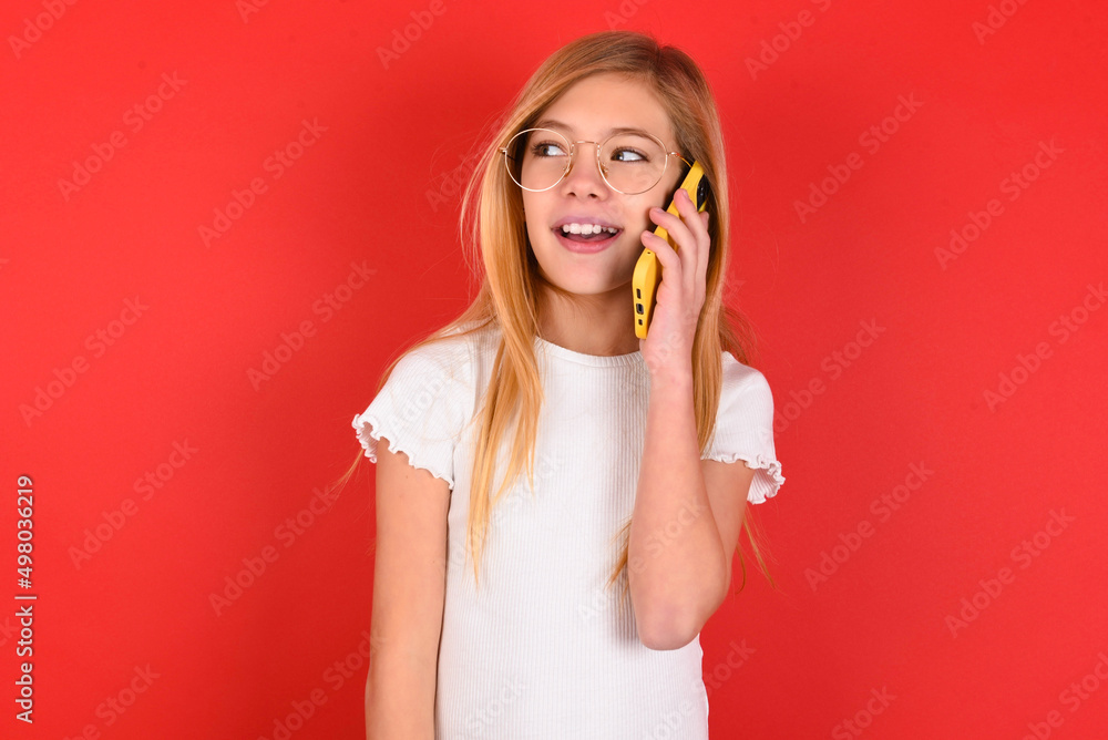 Pleasant looking happy blonde little kid girl wearing white t-shirt over red background has nice telephone conversation and looks aside, has nice mood and smiles positively while talks via cell phone Pleasant looking happy blonde little kid girl wearing white t-shirt over red background has nice telephone conversation and looks aside, has nice mood and smiles positively while talks via cell phone