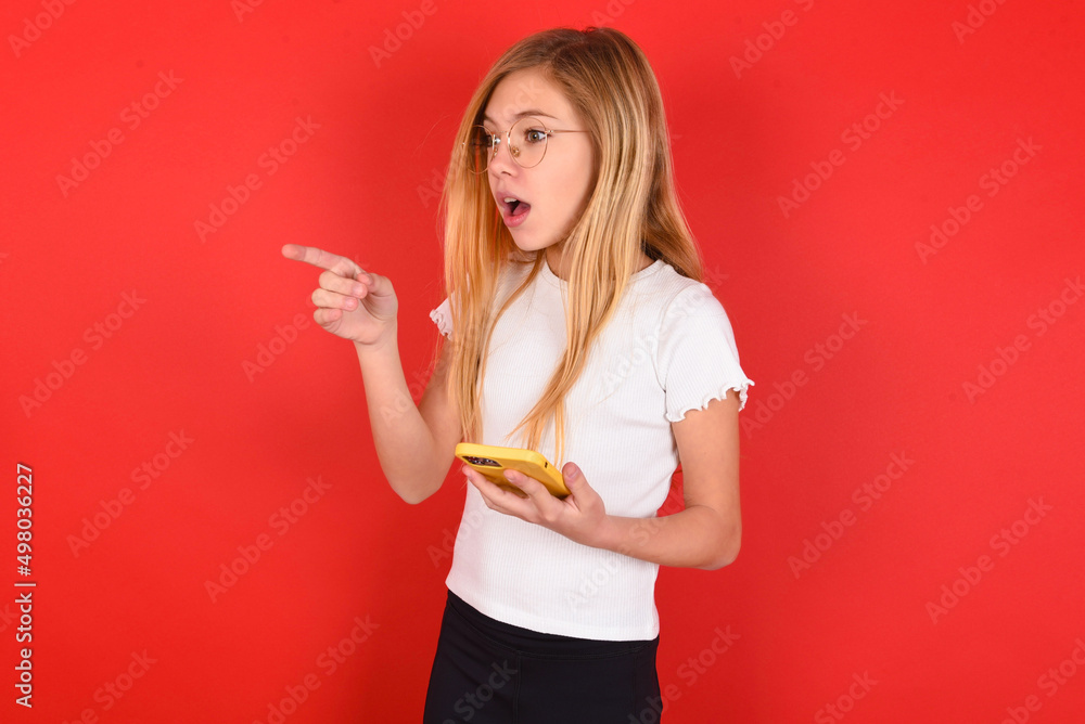 Stunned blonde little kid girl wearing white t-shirt over red background points sideways right copy space, recommends product, sees astonishing thing Stunned blonde little kid girl wearing white t-shirt over red background points sideways right copy space, recommends product, sees astonishing thing