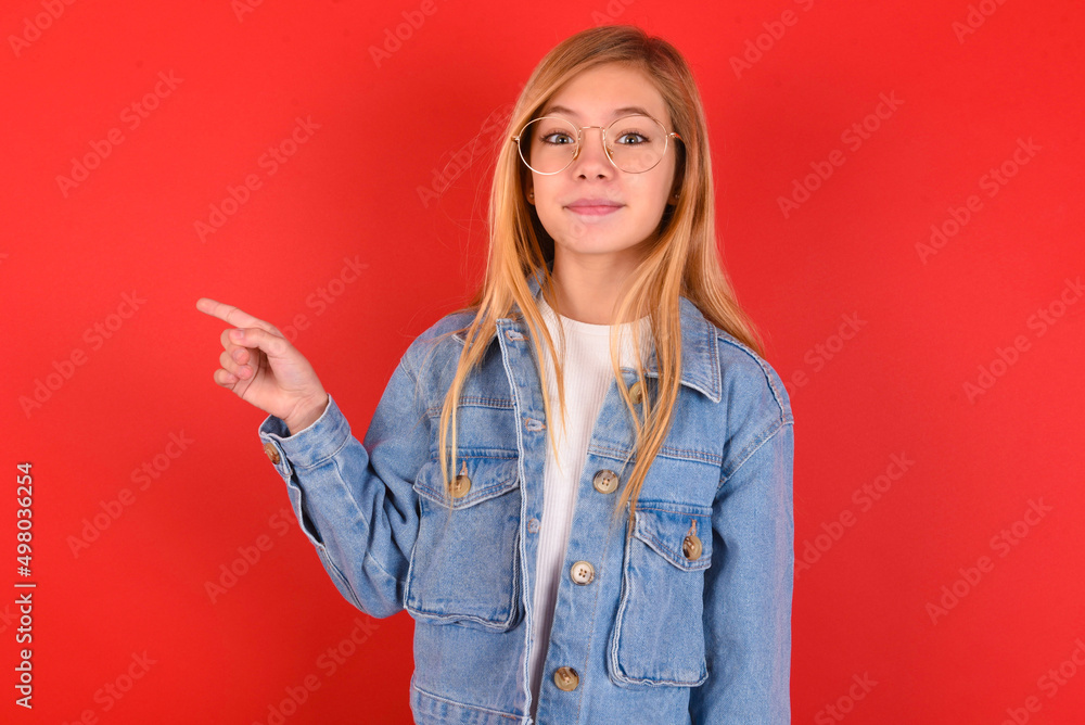 Positive blonde little kid girl wearing denim jacket over red background with satisfied expression indicates at upper right corner shows good offer suggests to click on link Positive blonde little kid girl wearing denim jacket over red background with satisfied expression indicates at upper right corner shows good offer suggests to click on link