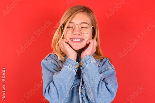 blonde little kid girl wearing denim jacket over red background grins joyfully, imagines something pleasant, copy space. Pleasant emotions concept.