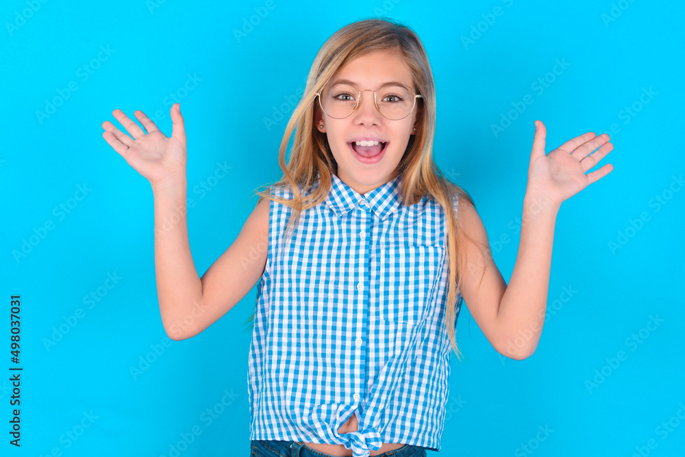 little kid girl with glasses wearing plaid shirt over blue background raising hands up, having eyes full of happiness rejoicing his great achievements. Achievement, success concept. little kid girl with glasses wearing plaid shirt over blue background raising hands up, having eyes full of happiness rejoicing his great achievements. Achievement, success concept.
