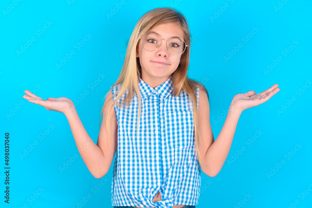 Maybe yes or no. Doubtful little kid girl with glasses wearing plaid shirt over blue wall shrugs shoulders in bewilderment, tries to make decision puzzled what he wants. Hesitation and uncertainty. Maybe yes or no. Doubtful little kid girl with glasses wearing plaid shirt over blue wall shrugs shoulders in bewilderment, tries to make decision puzzled what he wants. Hesitation and uncertainty.