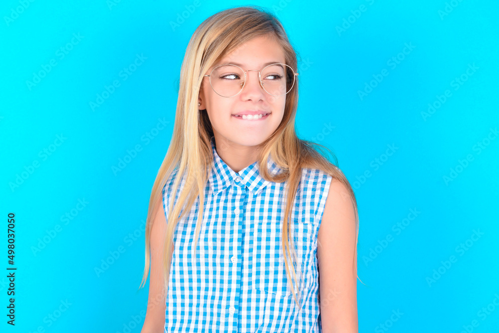 little kid girl with glasses wearing plaid shirt over blue background with thoughtful expression, looks away keeps hands down bitting his lip thinks about something pleasant. little kid girl with glasses wearing plaid shirt over blue background with thoughtful expression, looks away keeps hands down bitting his lip thinks about something pleasant.
