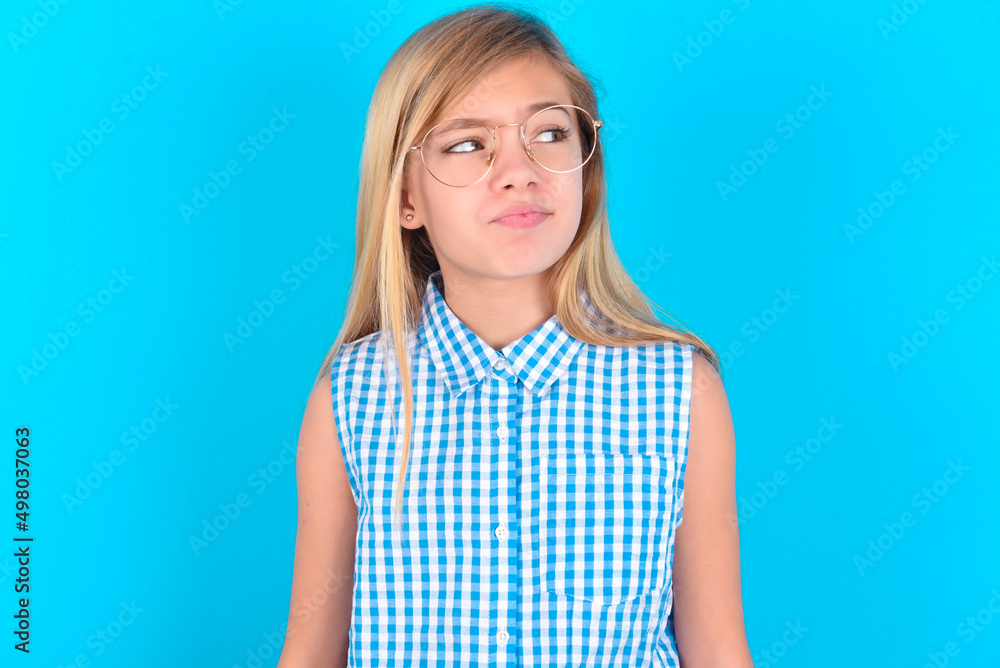 little kid girl with glasses wearing plaid shirt over blue background , looks pensively aside, plans actions after university, imagines what to do Thinks over about new project. little kid girl with glasses wearing plaid shirt over blue background , looks pensively aside, plans actions after university, imagines what to do Thinks over about new project.