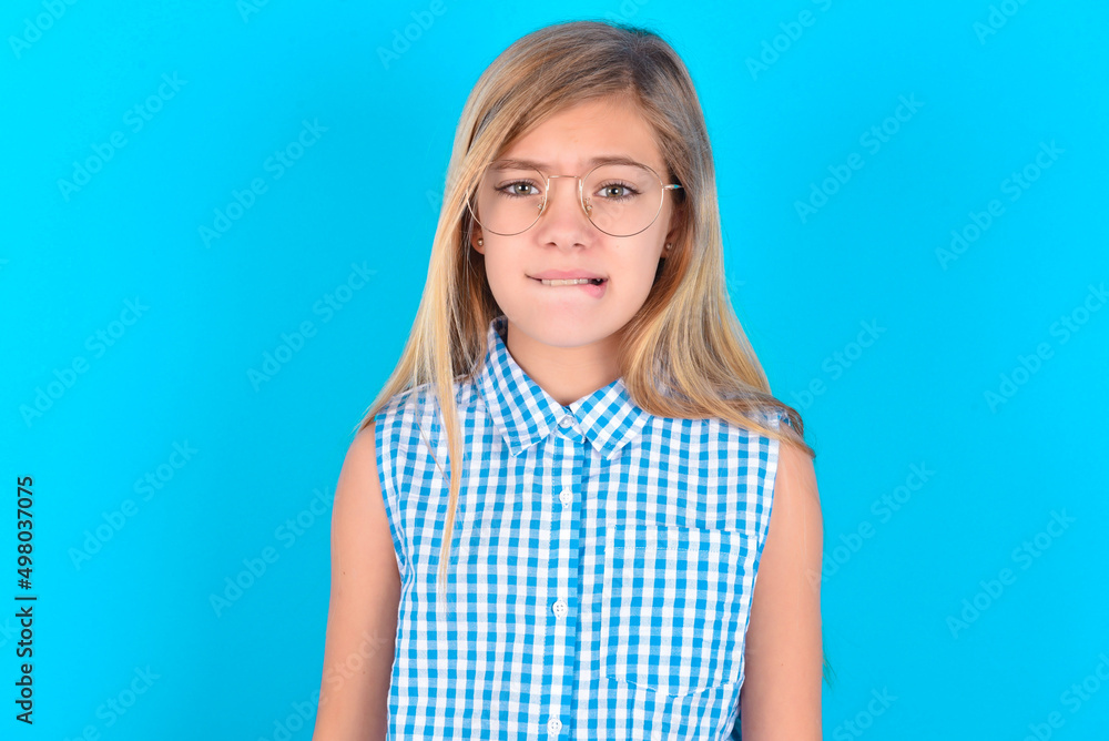little kid girl with glasses wearing plaid shirt over blue background being nervous and scared biting lips looking camera with impatient expression, pensive. little kid girl with glasses wearing plaid shirt over blue background being nervous and scared biting lips looking camera with impatient expression, pensive.