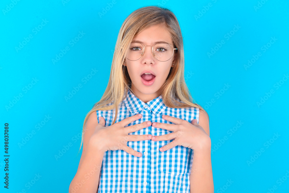 little kid girl with glasses wearing plaid shirt over blue background keeps hands on chest feeling shocked and scared, mouth widely opened, stares at camera saying: Who, me? little kid girl with glasses wearing plaid shirt over blue background keeps hands on chest feeling shocked and scared, mouth widely opened, stares at camera saying: Who, me?