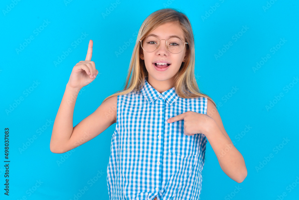 little kid girl with glasses wearing plaid shirt over blue background says: wow how exciting it is, has amazed expression, indicates something. One hand on her chest and pointing with other hand. little kid girl with glasses wearing plaid shirt over blue background says: wow how exciting it is, has amazed expression, indicates something. One hand on her chest and pointing with other hand.