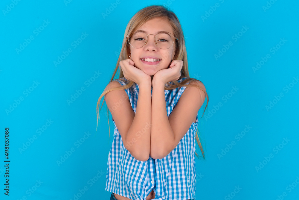 Dreamy little kid girl with glasses wearing plaid shirt over blue background keeps hands pressed together under chin, looks with happy expression, has toothy smile. Dreamy little kid girl with glasses wearing plaid shirt over blue background keeps hands pressed together under chin, looks with happy expression, has toothy smile.