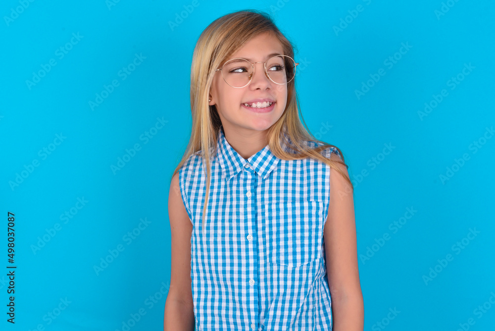 Oops! Portrait of little kid girl with glasses wearing plaid shirt over blue background clenches teeth and looks confusedly aside, realizes her bad mistake, Oops! Portrait of little kid girl with glasses wearing plaid shirt over blue background clenches teeth and looks confusedly aside, realizes her bad mistake,