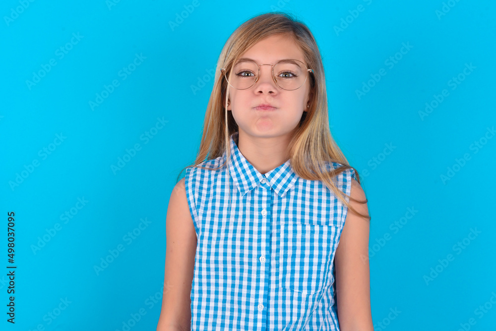 little kid girl with glasses wearing plaid shirt over blue background  puffing cheeks with funny face. Mouth inflated with air, crazy expression. - obrazy, fototapety, plakaty 