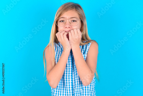 Anxiety - little kid girl with glasses wearing plaid shirt over blue background covering his mouth with hands scared from something or someone bitting nails.