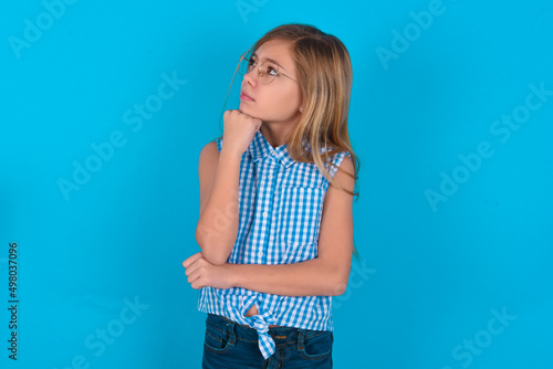 Portrait of thoughtful little kid girl with glasses wearing plaid shirt over blue back keeps hand under chin, looks away trying to remember something or listens something with interest. Youth concept.