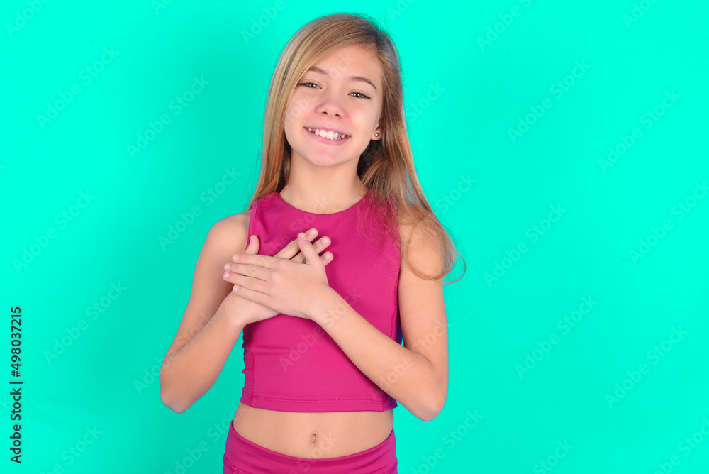 Honest blonde little kid girl wearing pink sport clothes over green background keeps hands on chest, touched by compliment or makes promise, looks at camera with great pleasure. Honest blonde little kid girl wearing pink sport clothes over green background keeps hands on chest, touched by compliment or makes promise, looks at camera with great pleasure.