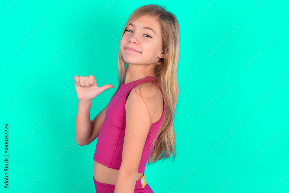 Closeup of cheerful blonde little kid girl wearing pink sport clothes over green background looks joyful, satisfied and confident, points at himself with thumb. Closeup of cheerful blonde little kid girl wearing pink sport clothes over green background looks joyful, satisfied and confident, points at himself with thumb.
