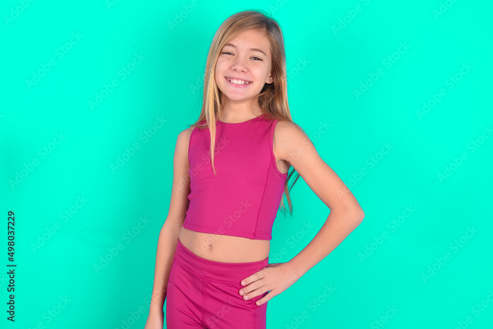 Studio shot of cheerful blonde little kid girl wearing pink sport clothes over green background keeps hand on hip, smiles broadly. Studio shot of cheerful blonde little kid girl wearing pink sport clothes over green background keeps hand on hip, smiles broadly.