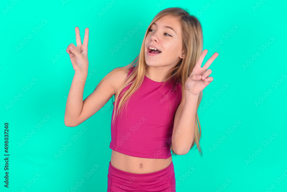 Isolated shot of cheerful blonde little kid girl wearing pink sport clothes over green background makes peace or victory sign with both hands, feels cool. Isolated shot of cheerful blonde little kid girl wearing pink sport clothes over green background makes peace or victory sign with both hands, feels cool.