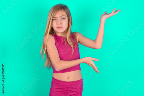 blonde little kid girl wearing pink sport clothes over green background pointing aside with both hands showing something strange and saying: I don't know what is this. Advertisement concept. photo
