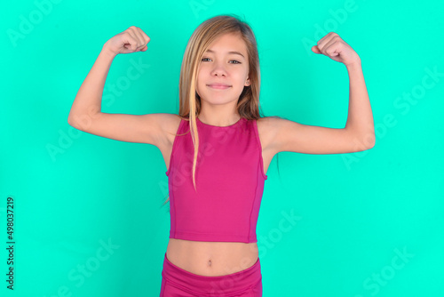 Waist up shot of blonde little kid girl wearing pink sport clothes raises arms to show muscles feels confident in victory, looks strong and independent, smiles positively at camera. Sport concept. © Jihan