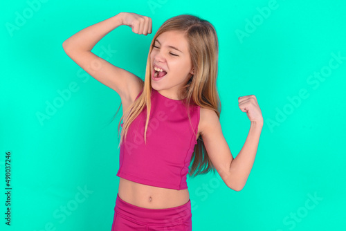 Yes I am winner. Portrait of charming delighted and excited blonde little kid girl wearing pink sport clothes raising up fist in triumph and victory smiling achieving success grinning from delight. © Jihan