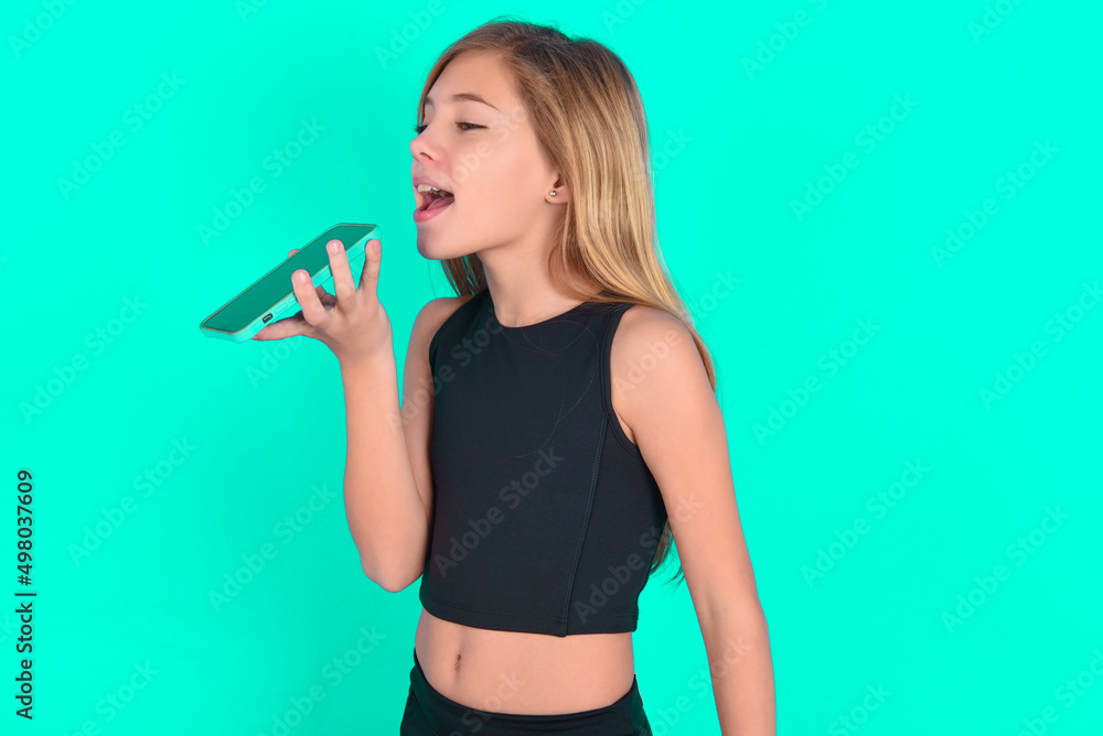 Smiling blonde little kid girl wearing black sport clothes over green background sending voice message on her smart phone. Communication and new technologies concept.