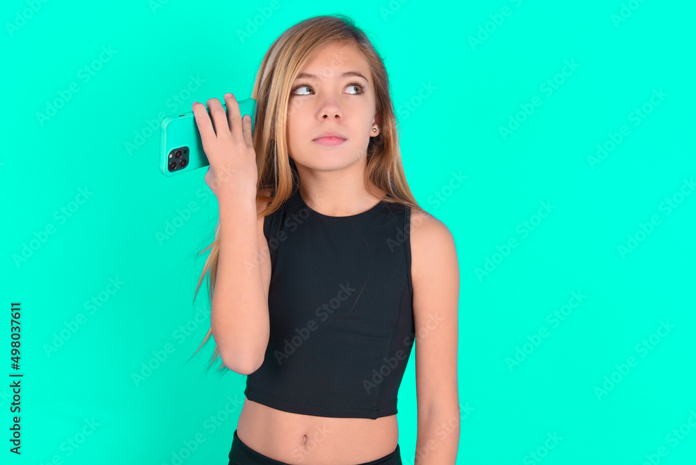 Smiling blonde little kid girl wearing black sport clothes over green background listening a voice message from her smartphone. Communication and technology concept. Smiling blonde little kid girl wearing black sport clothes over green background listening a voice message from her smartphone. Communication and technology concept.