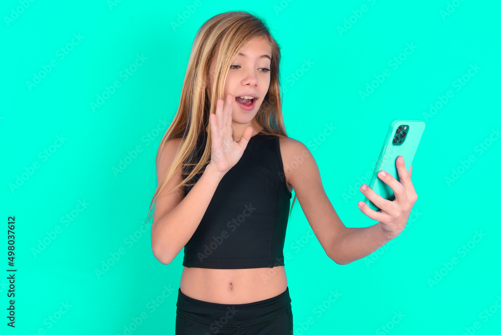 Portrait of happy friendly blonde little kid girl wearing black sport clothes over green background taking selfie and waving hand, communicating on video call, online chatting. Portrait of happy friendly blonde little kid girl wearing black sport clothes over green background taking selfie and waving hand, communicating on video call, online chatting.